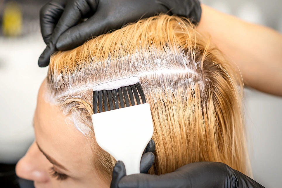 A person wearing gloves applies hair dye with a brush to a blonde-haired woman's roots in a salon.