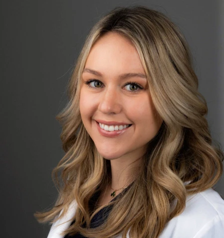 A young woman with long, wavy blonde hair is smiling and looking at the camera. She is wearing a white coat and a dark top. The background is plain and gray.