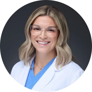 A woman with shoulder-length blonde hair, wearing eyeglasses, a blue top, and a white lab coat, smiles in this professional headshot.