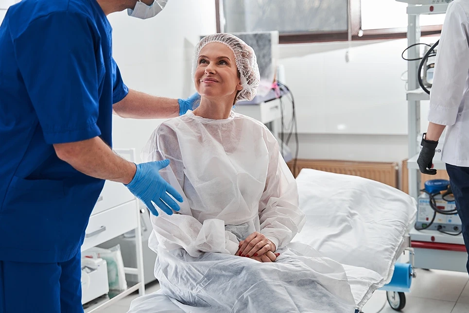 A person in protective clothing sits on a medical examination table, while a healthcare professional in blue scrubs and gloves talks to them. Another person is in the background.