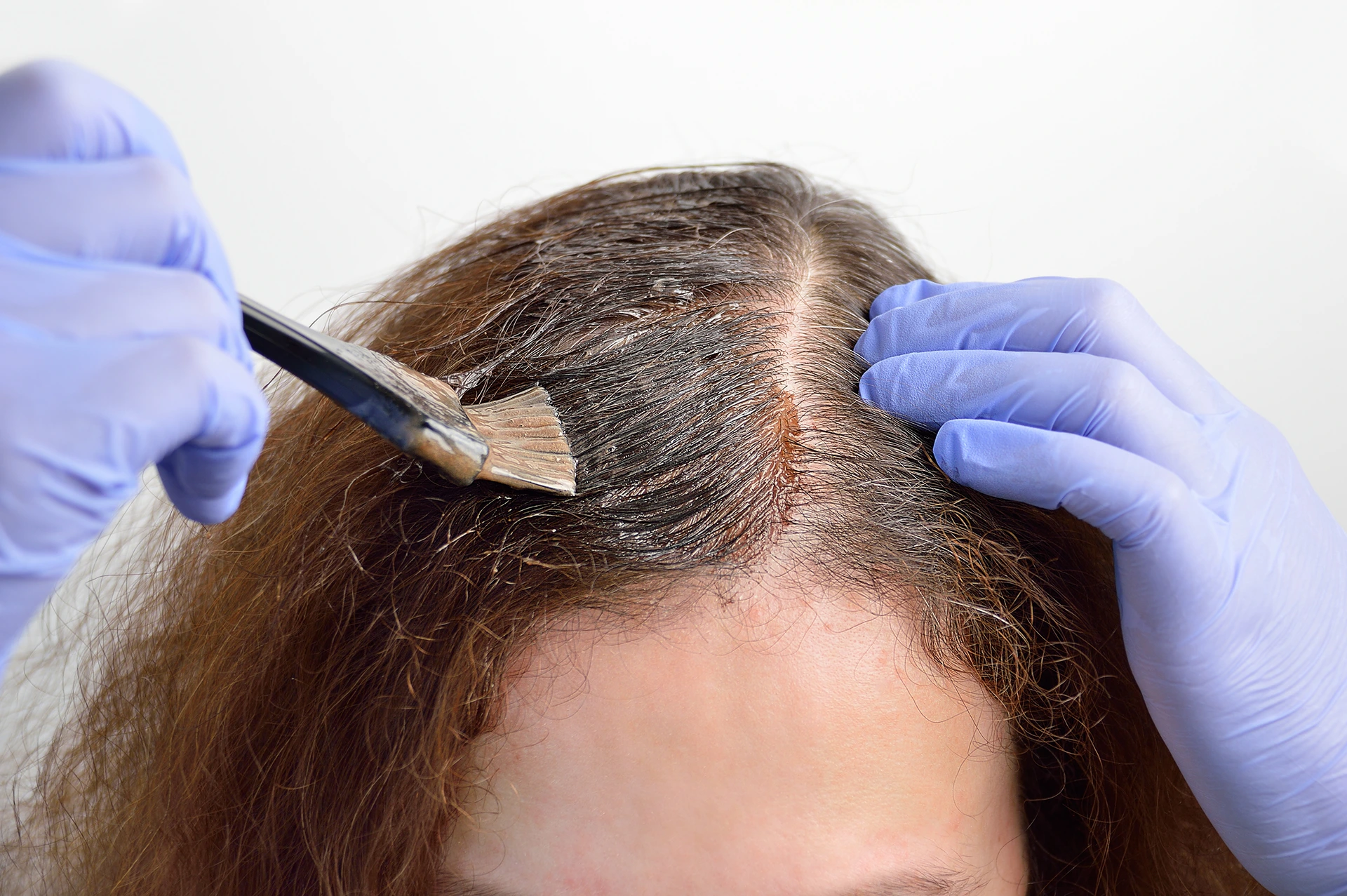 Two hands wearing blue gloves applying hair dye to a woman's brown hair with a brush, focusing on the scalp area.
