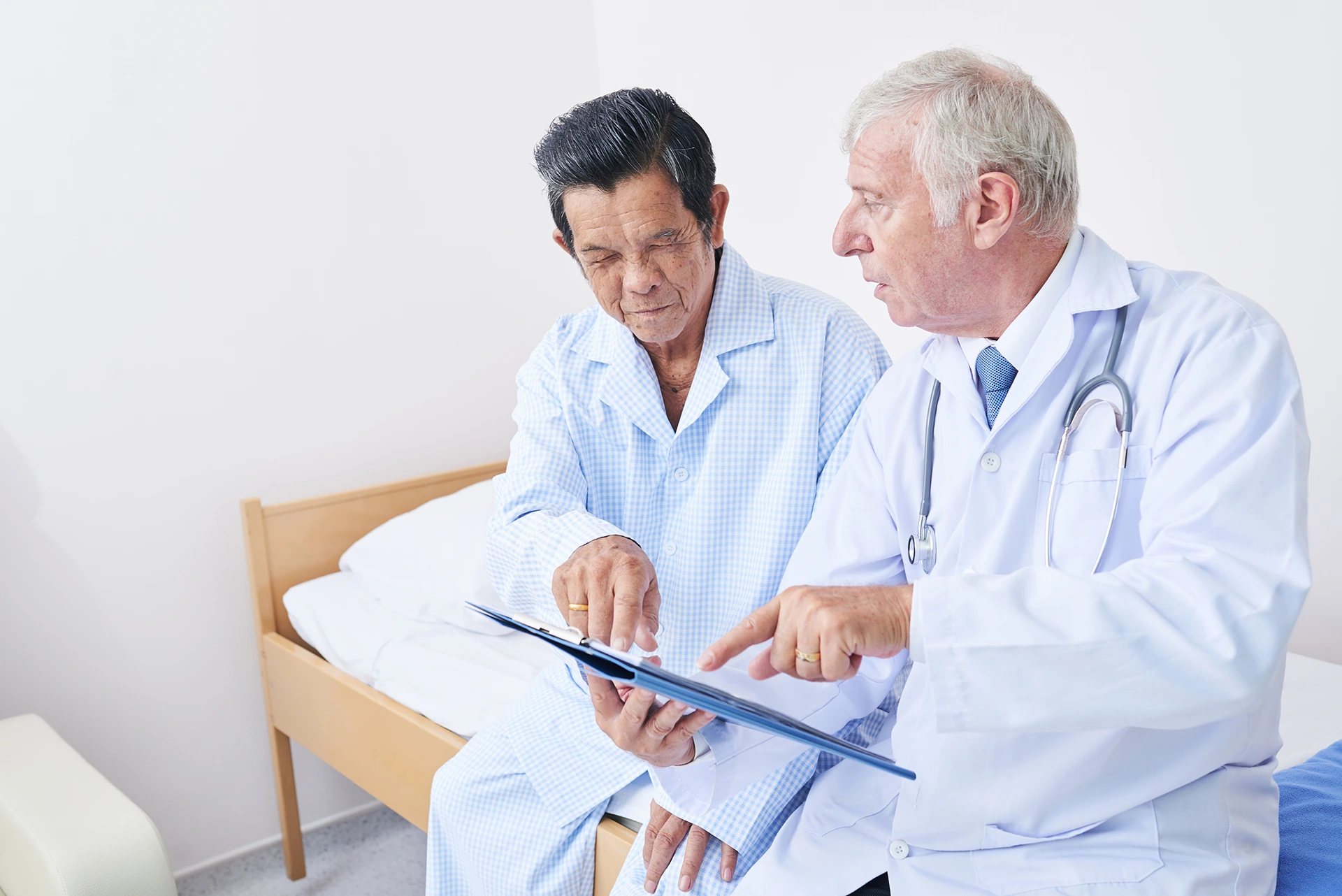 A doctor in a white coat discusses a document with an elderly male patient sitting on a bed in a medical gown. Both are focused on the document.