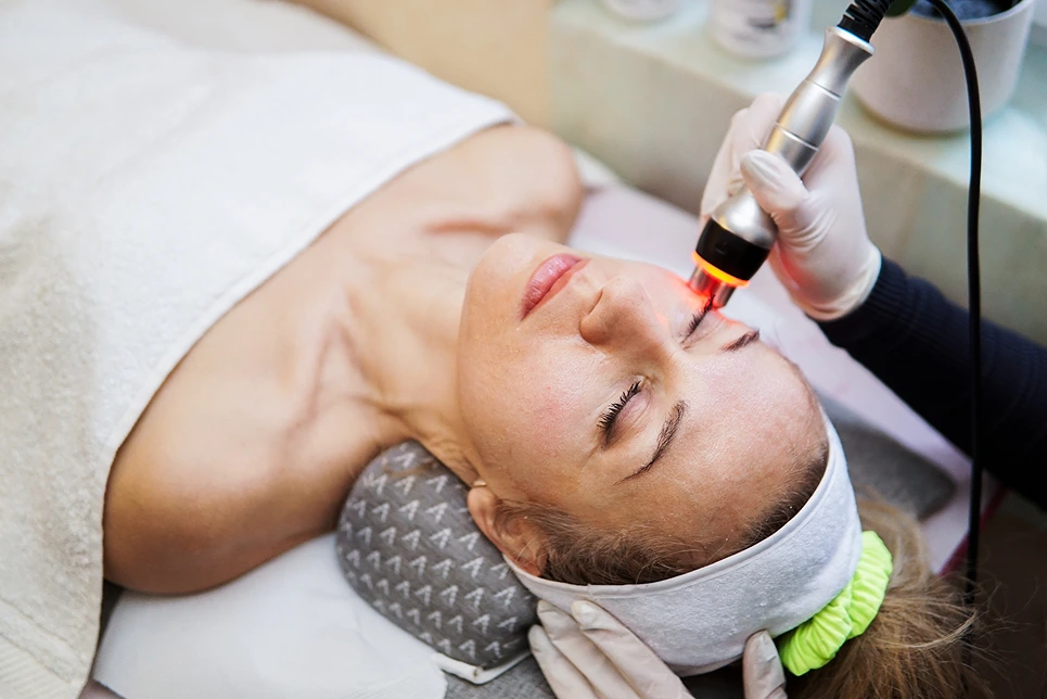 A woman is lying on a treatment table with a headband, receiving a laser facial treatment from a professional wearing gloves.