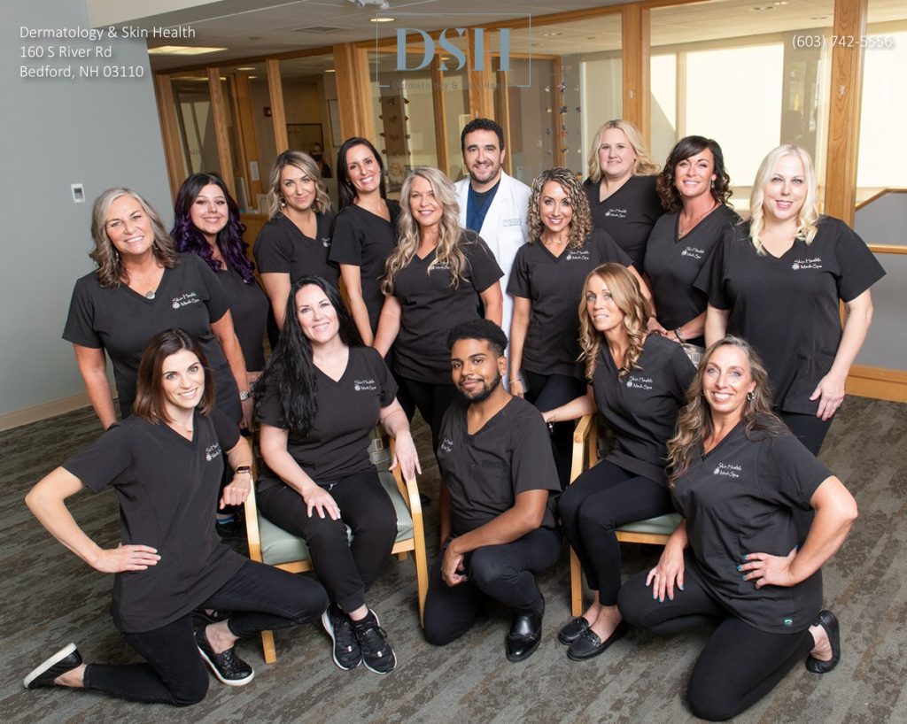 A group of people in black uniforms and a person in a white coat pose indoors at Dermatology & Skin Health in Bedford, NH. The group is smiling and standing or sitting in two rows.