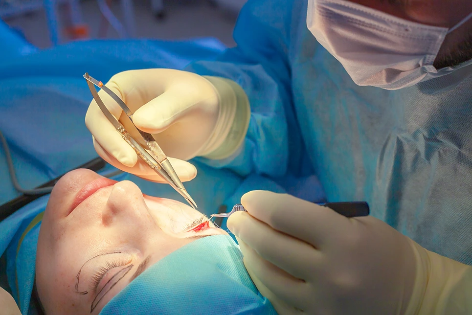 A surgeon performs a delicate surgical procedure on a patient's face, using a pair of forceps and a scalpel. Both are dressed in surgical attire and gloves.