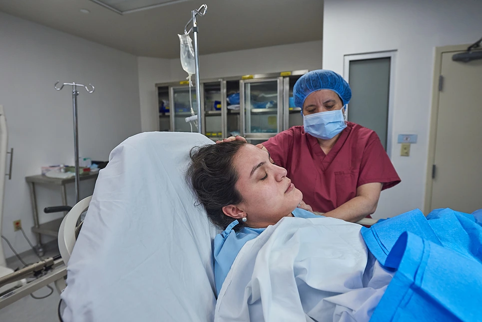 A patient lying in a hospital bed with an IV line, while a healthcare worker in scrubs and a hair net stands beside her, adjusting the blanket.