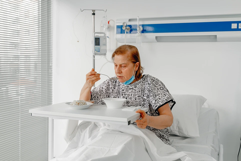 A patient in a hospital bed is eating a meal placed on a tray table. Medical equipment is visible in the background.