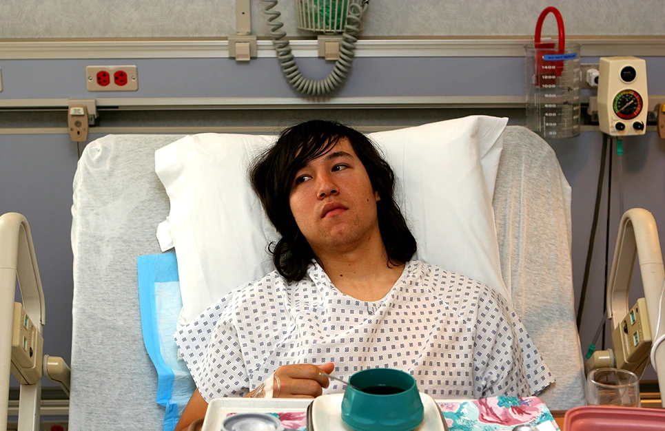 Person in a hospital bed wearing a hospital gown with medical equipment in the background, holding a mug.