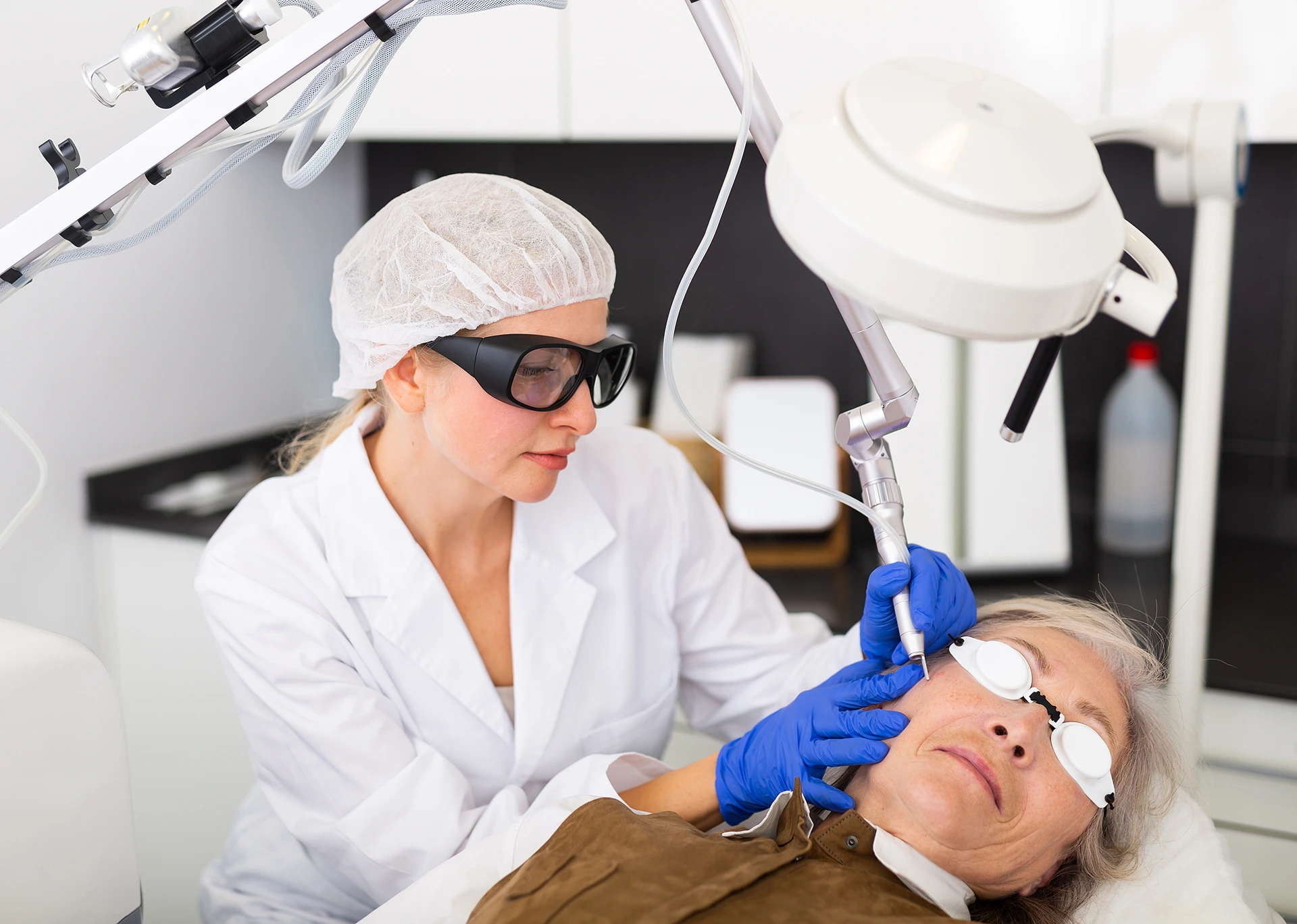 A medical professional wearing protective gear performs a laser procedure on an older patient lying down, who is also wearing eye protection.