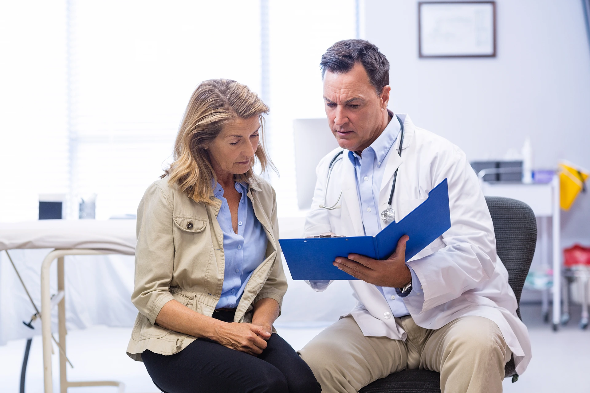 A doctor discusses medical information with a patient, both sitting in an examination room. The doctor is holding a blue clipboard and they appear to be reviewing documents.