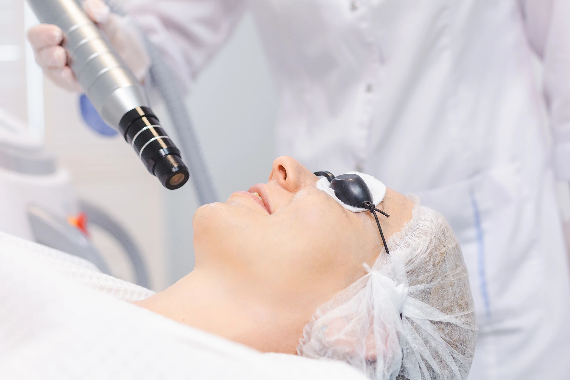 Person lying down undergoing a laser skin treatment, wearing protective goggles and surgical cap, with a practitioner holding a laser device near their face.