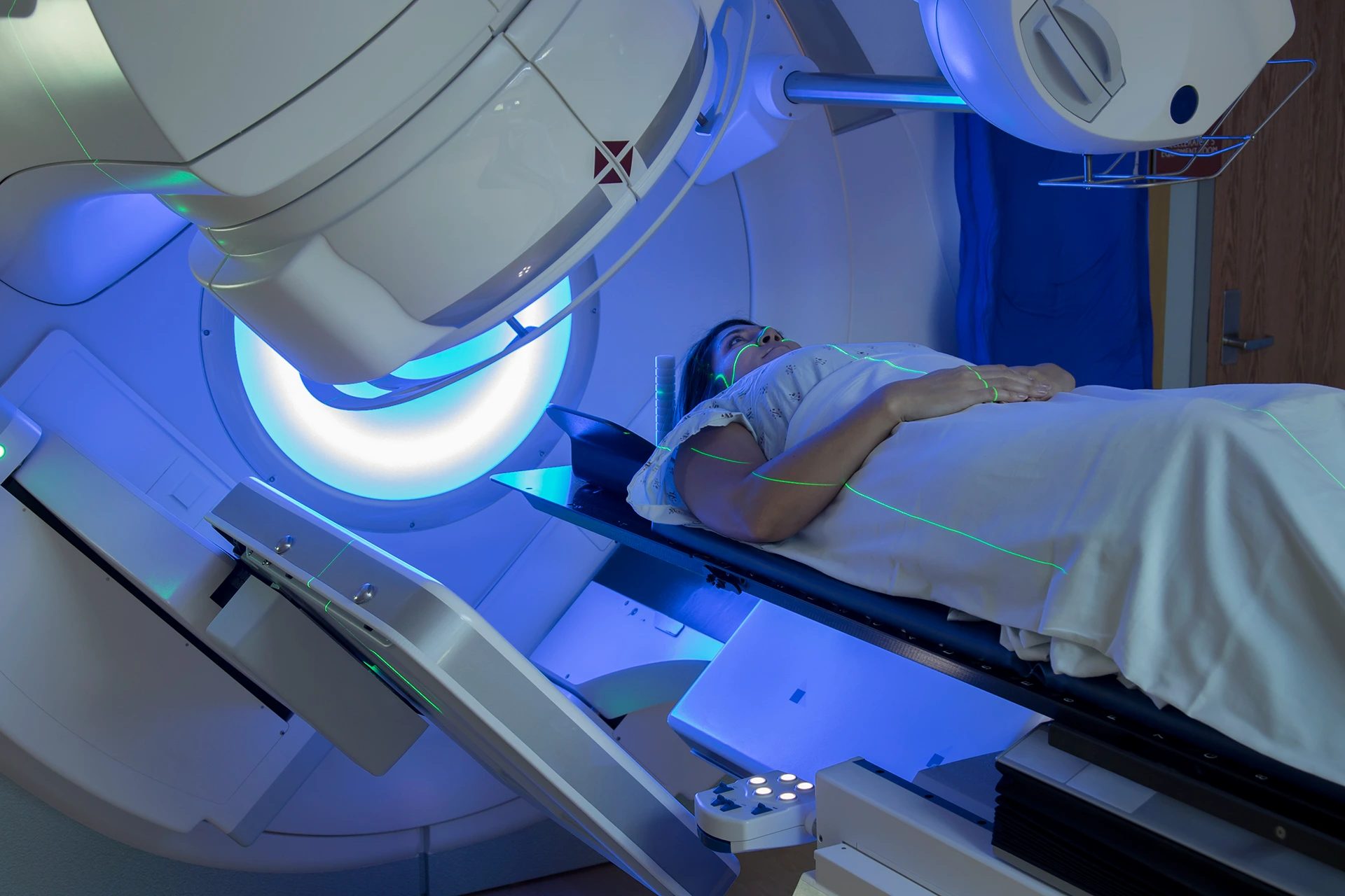 A person lies under a large machine undergoing a medical procedure, illuminated by blue light. The person is covered with a white sheet.