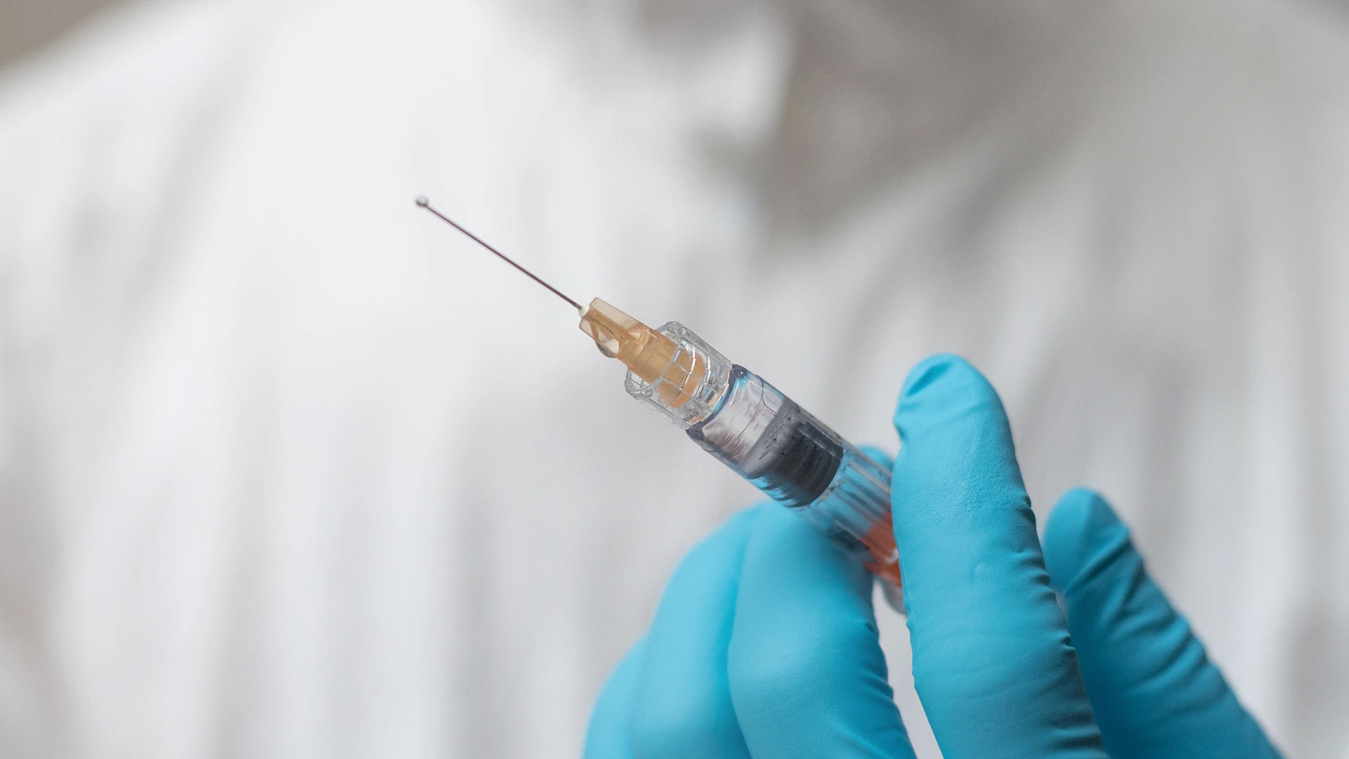 A gloved hand holds a syringe filled with liquid, ready for injection. The background shows a white, blurred surface.