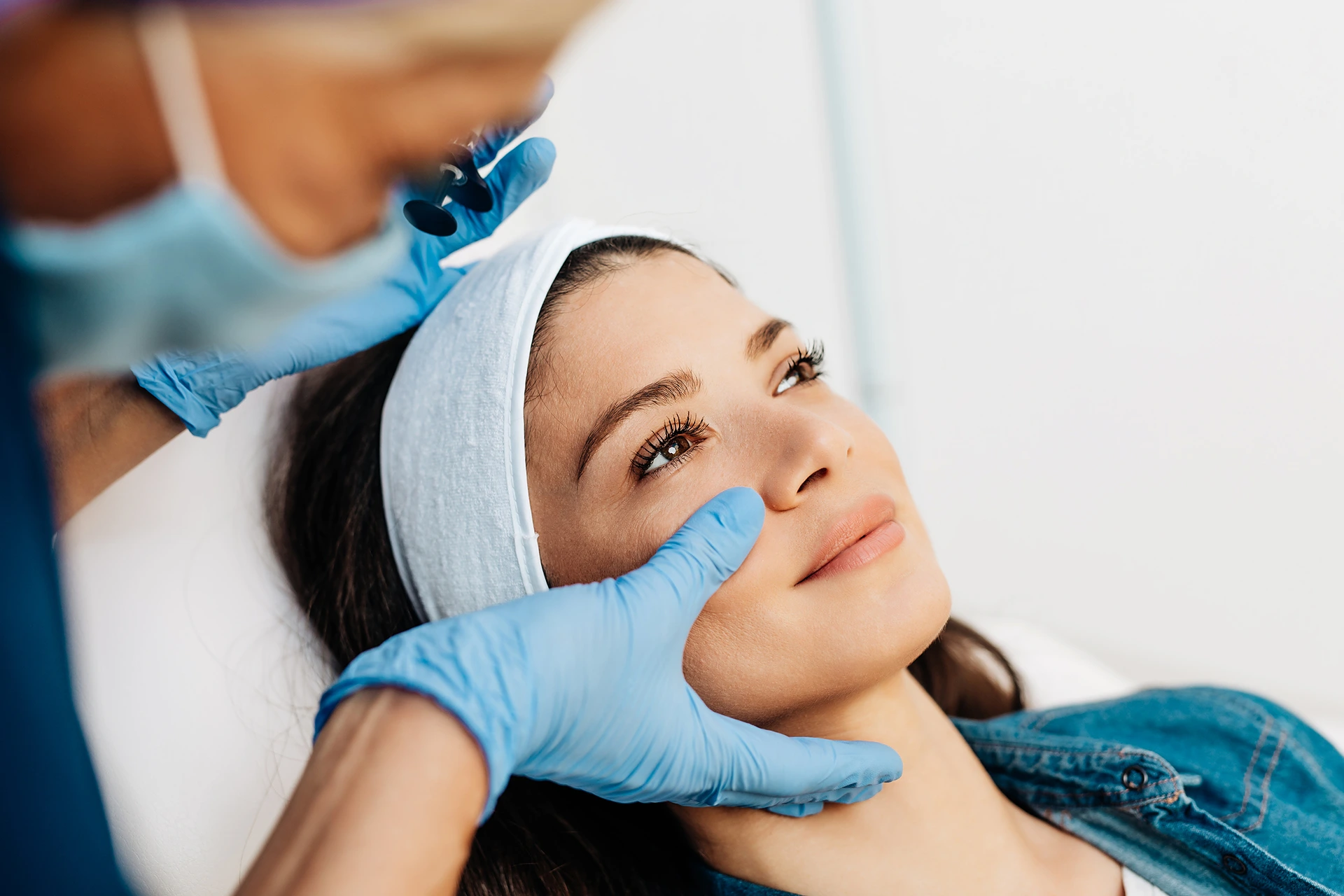 A woman with a headband lies back as a person wearing blue gloves performs a cosmetic procedure on her face.