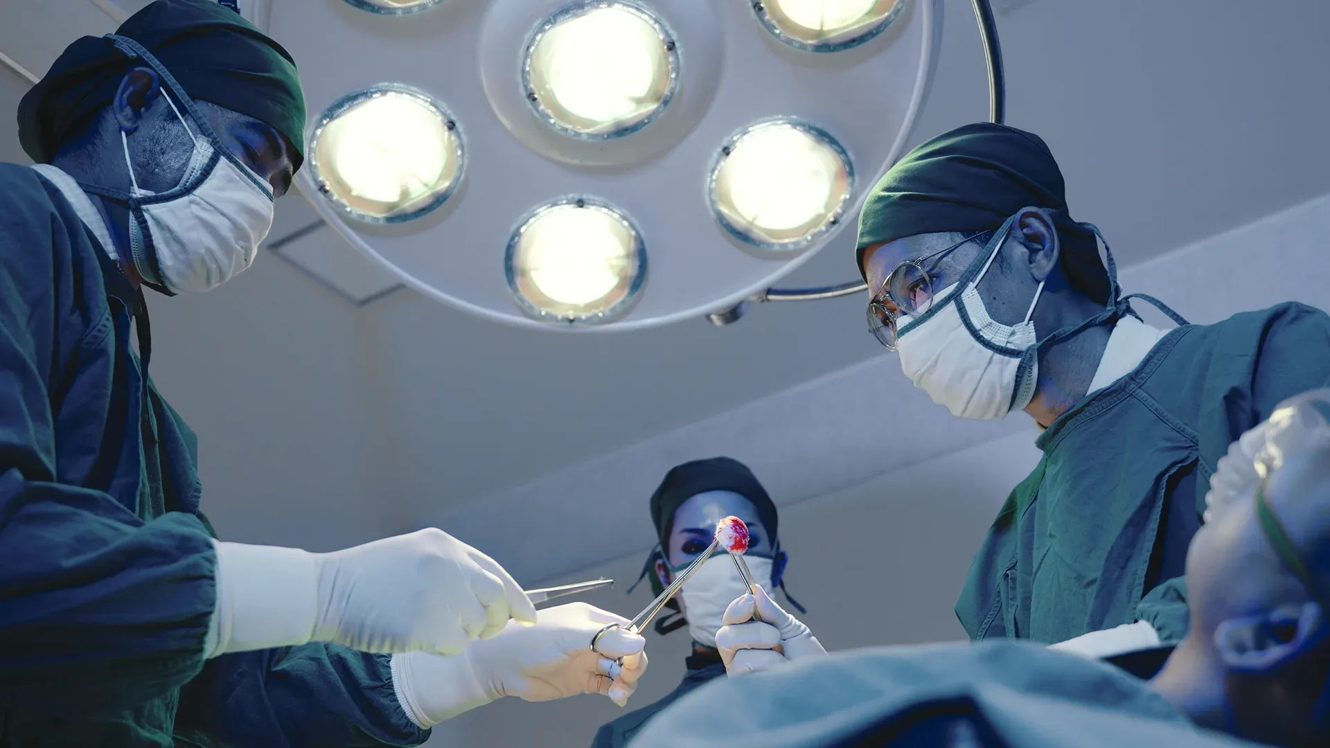 Surgeons in an operating room perform surgery under bright lights, all wearing green scrubs, masks, and gloves.