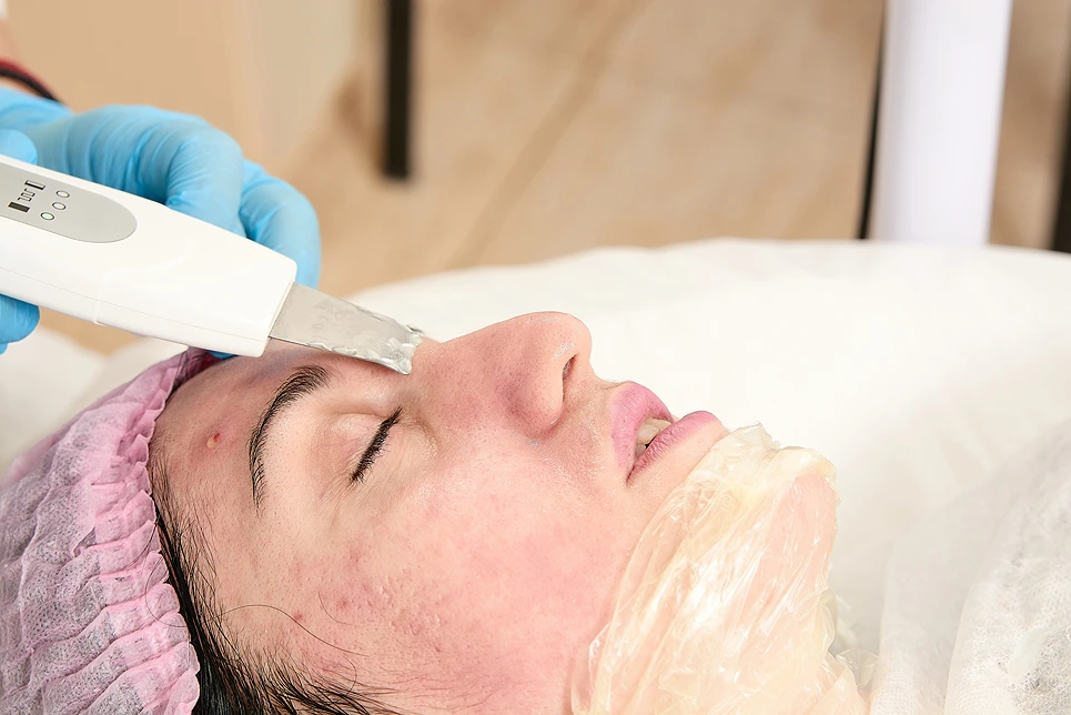 Close-up of a person receiving a facial treatment with a handheld device, while lying down with eyes closed and a pink hair cap.