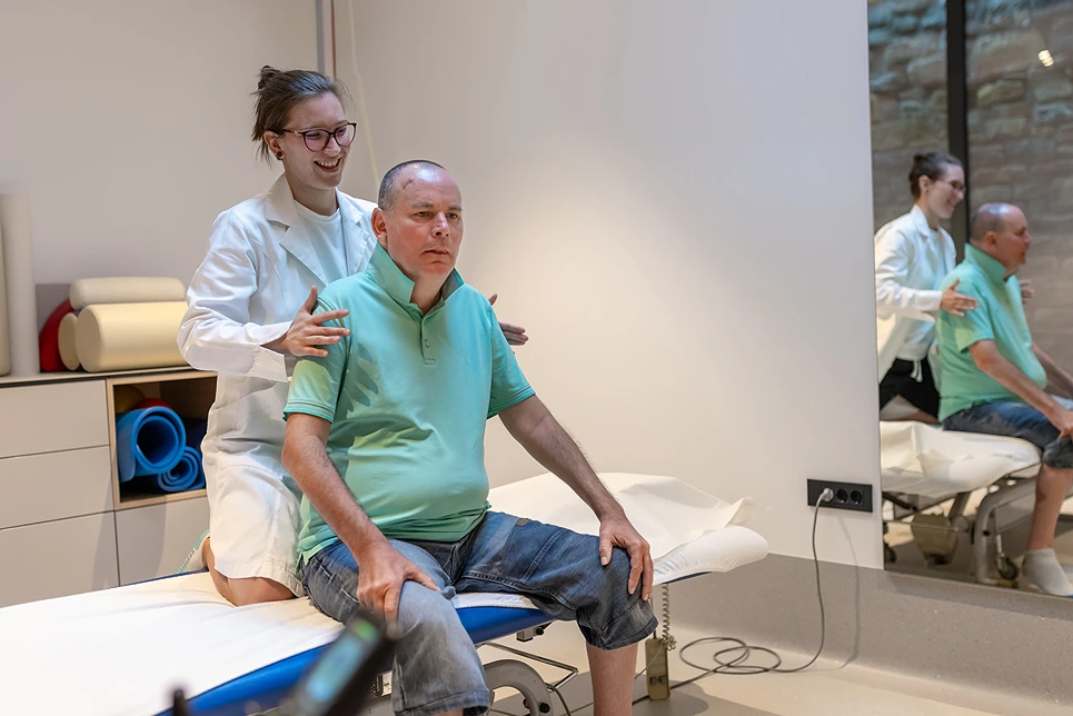 A physical therapist guides a man through a seated exercise in a modern clinical setting, with both facing forward.