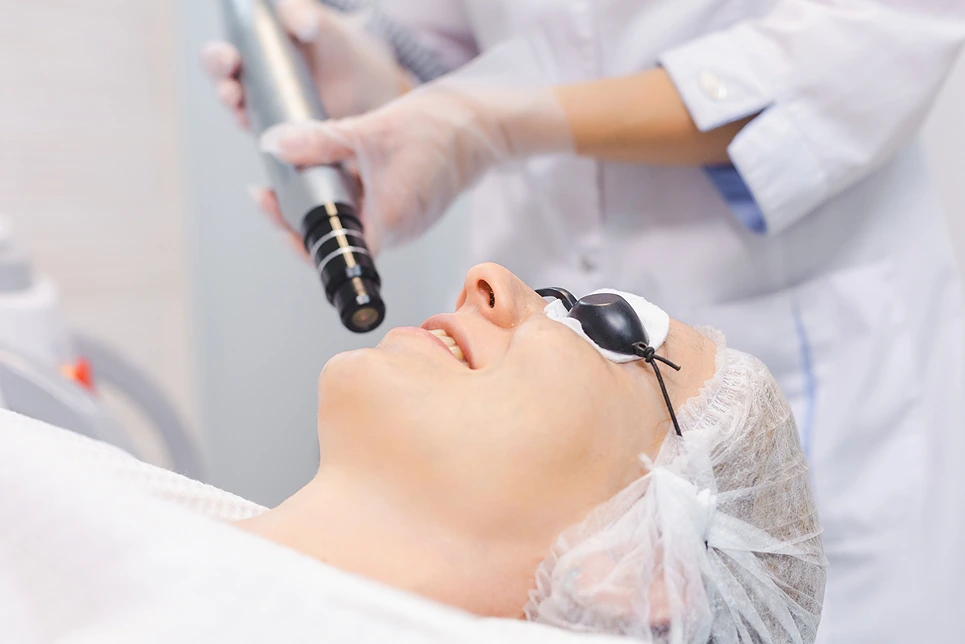 A person lies on a medical bed wearing protective eyewear while a healthcare professional in gloves and a lab coat holds a laser device above their face.