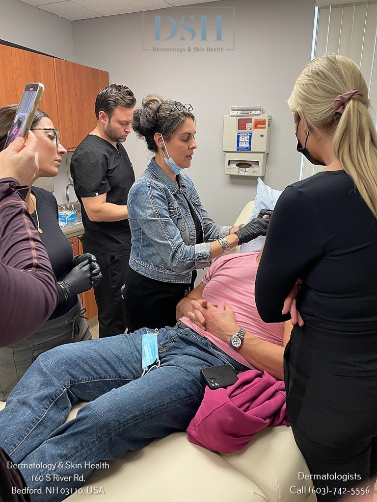 A medical professional injects a patient lying on an examination table, with three others observing in a dermatology clinic. Contact information for the clinic is displayed in the image.